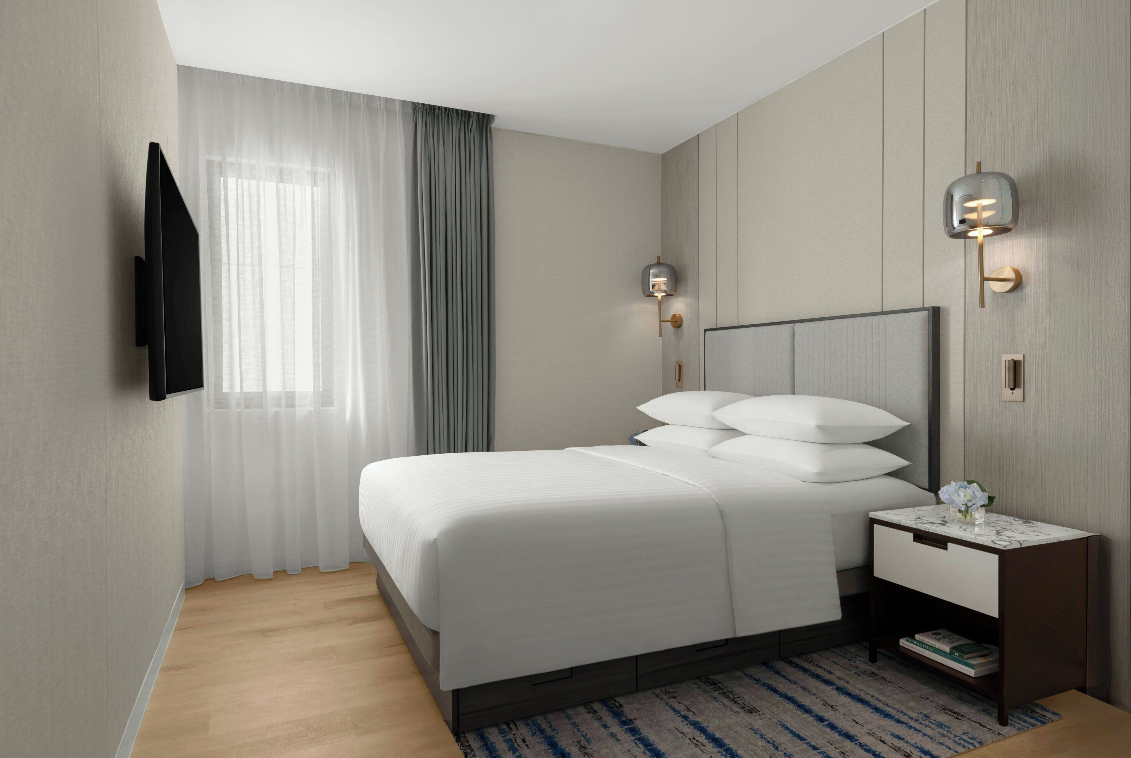 A modern hotel room with a large bed, a bedside table with a lamp, and a flat-screen TV mounted on the wall.