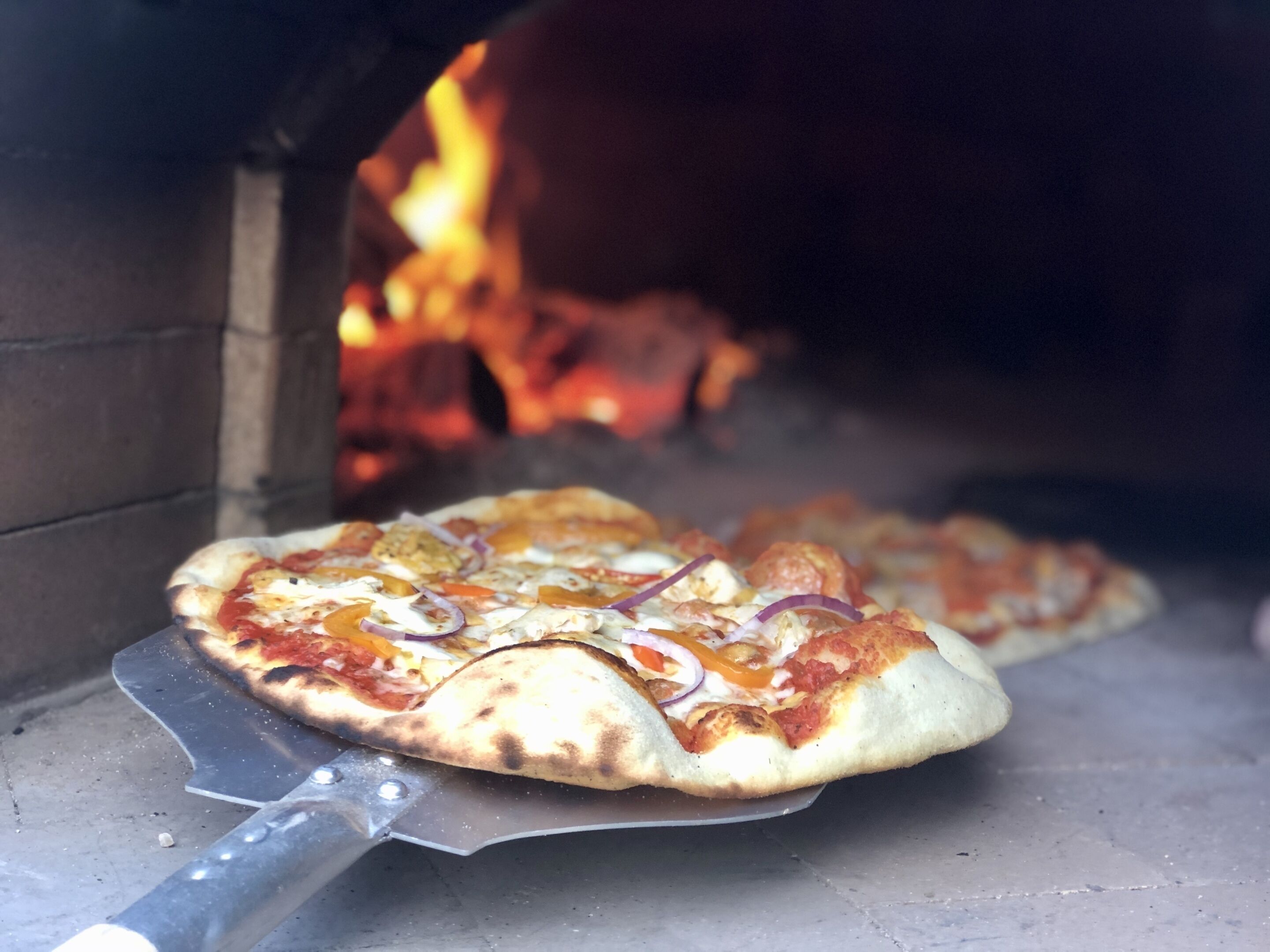Ein Pizzaofen mit einer Pizza darin, sichtbare Flammen und einem Pizzaschieber.