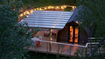 A wooden cabin with a sloped roof, a deck with a table and chairs, and a fire pit area.