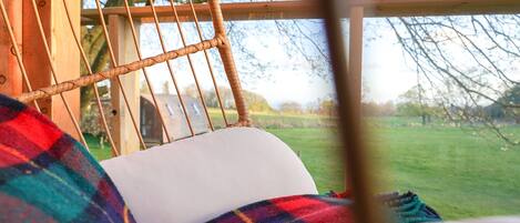 A wicker chair with a white cushion, a plaid blanket, and a view of a grassy field through a window.
