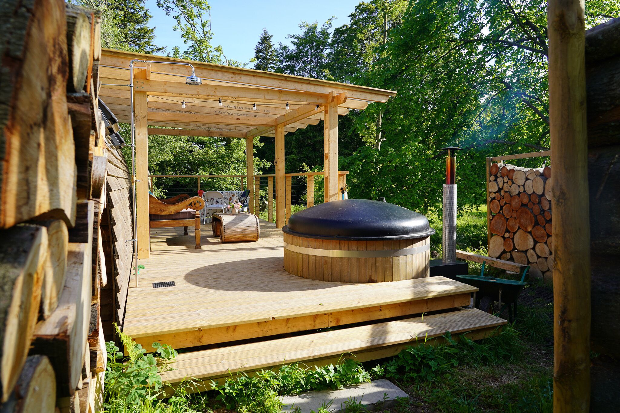 A wooden deck with a hot tub, a covered seating area, and a stack of firewood.