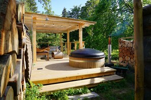 Une terrasse en bois avec un jacuzzi, un espace salon abrité et une pile de bois de chauffage.