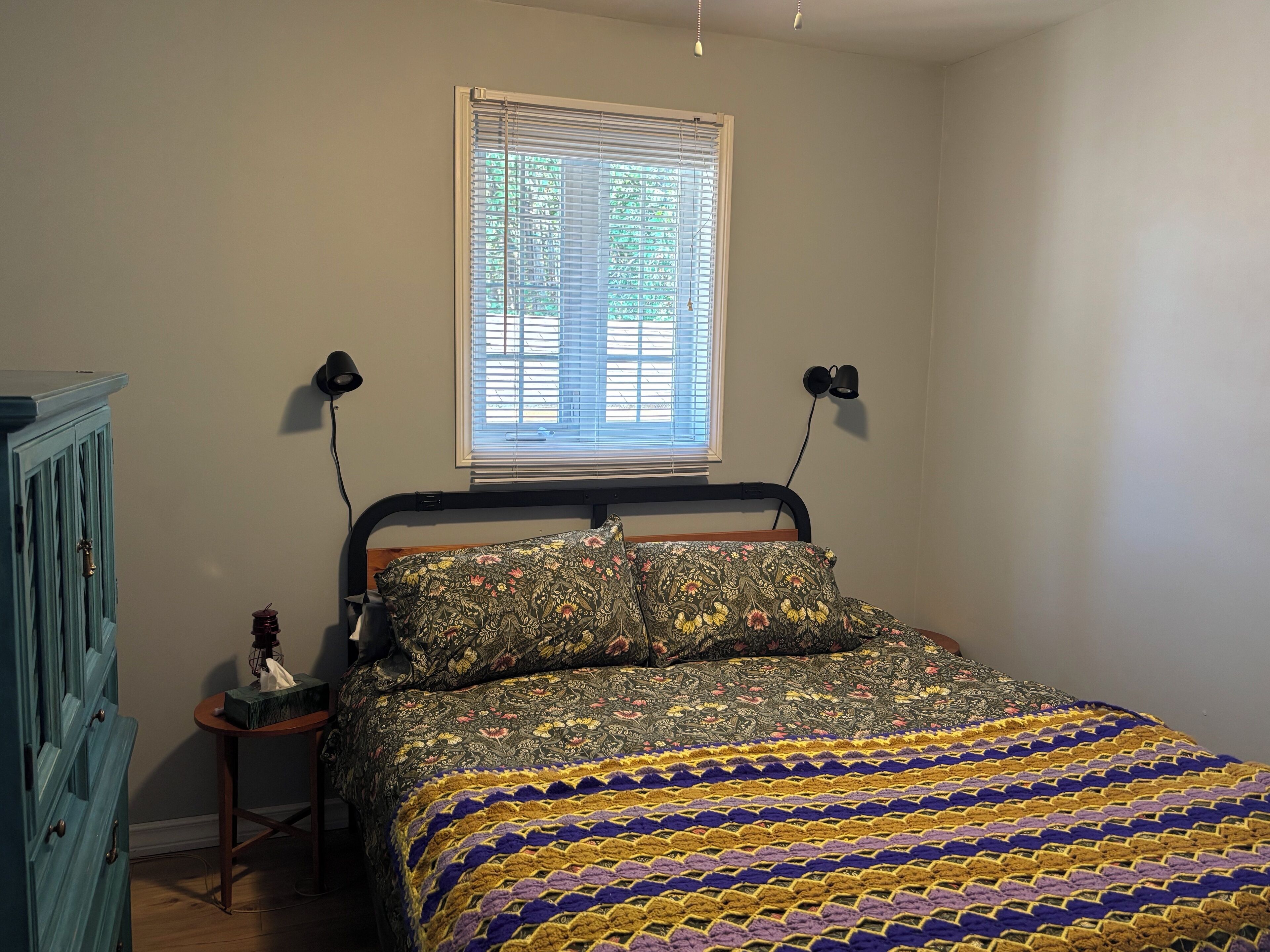 Un dormitorio con cama, mesita de noche, lámpara, ventana con persianas y un armario azul.