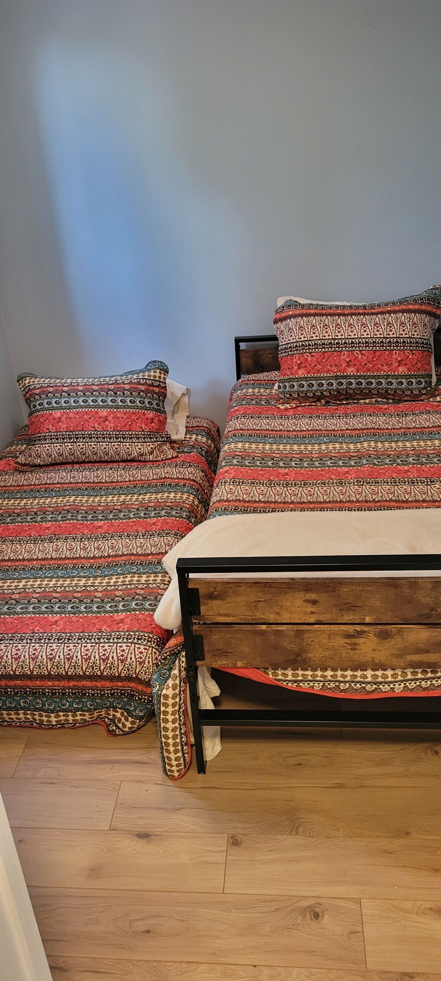 A bedroom with two beds, each with patterned bedding and pillows. The beds are made with a wooden frame and a red and beige blanket. The room has wooden flooring and a plain wall.