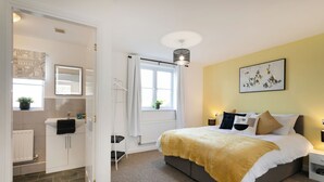 Un dormitorio con cama, mesitas de noche, ventana con cortinas, radiador, lavamanos y espejo en un baño.