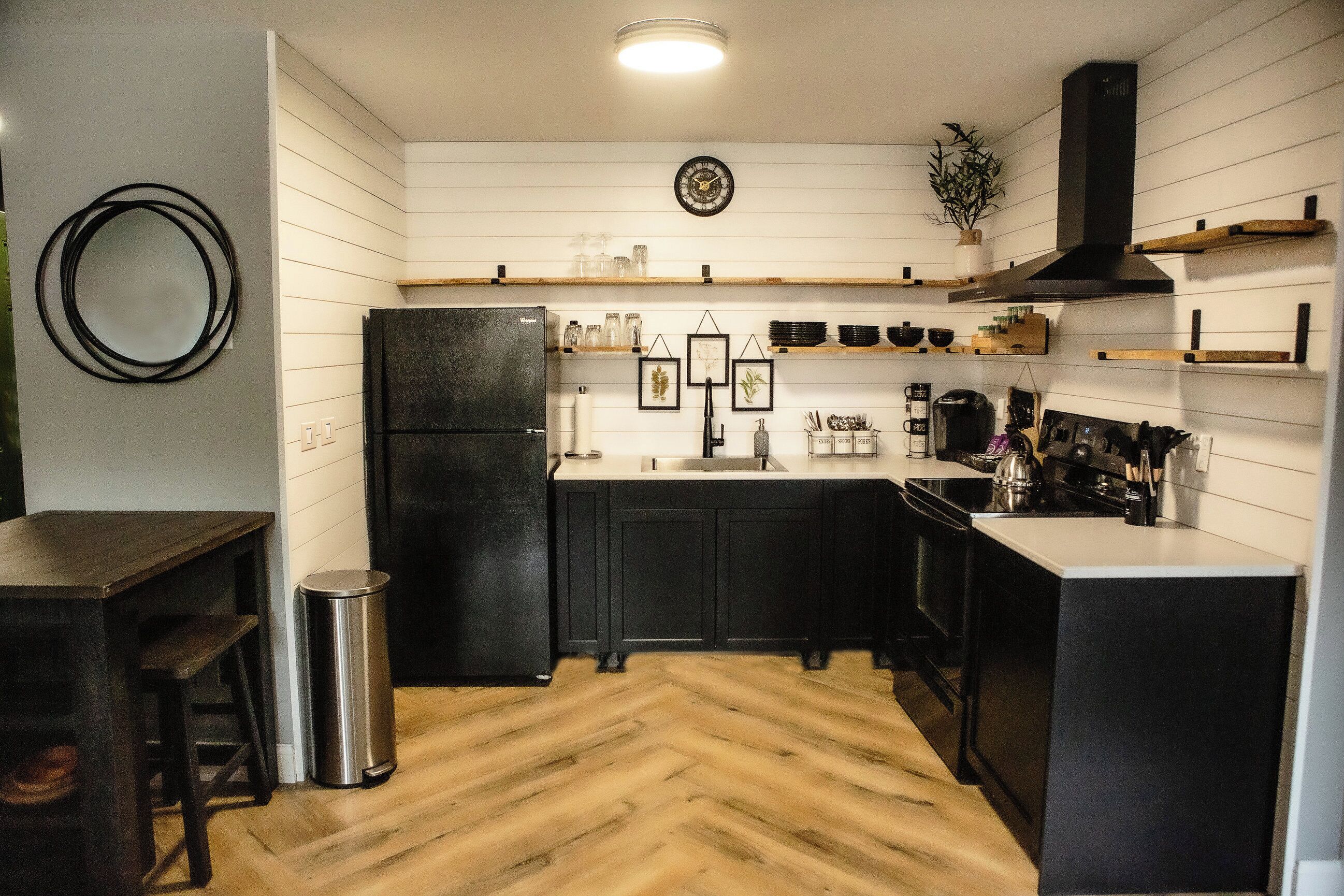 Een moderne keuken met witte panelen, een zwarte koelkast, houten vloeren en een roestvrijstalen prullenbak.