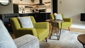 Ein Wohnzimmer mit zwei grünen Sesseln, einem runden Couchtisch und einer Küche im Hintergrund.