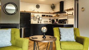 Ein modernes Wohnzimmer mit zwei grünen Sesseln, einem runden Couchtisch und einer Küchenzeile mit Holzregalen und einem schwarzen Herd.
