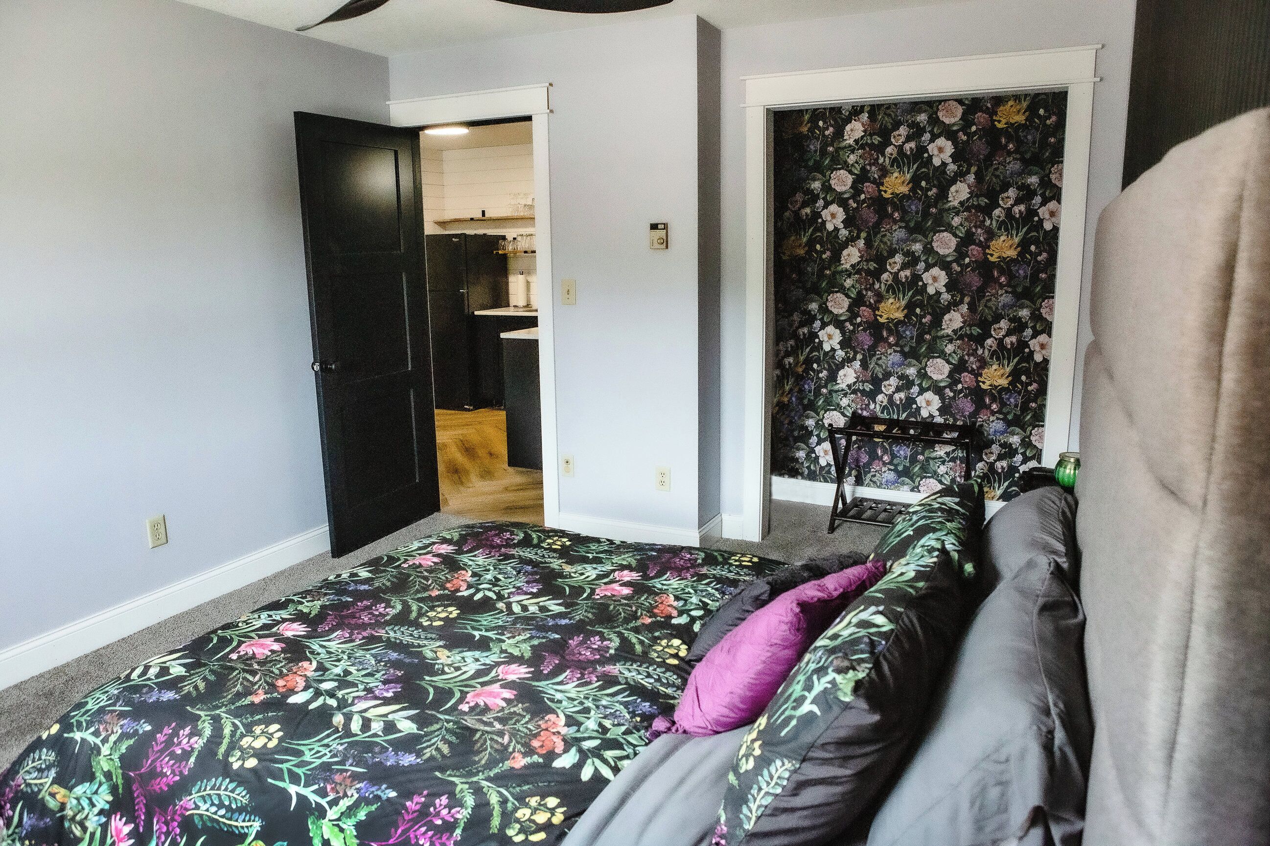 Ett sovrum med ett blommigt sängöverkast, en grå soffa och en dörr som leder in till ett kök.