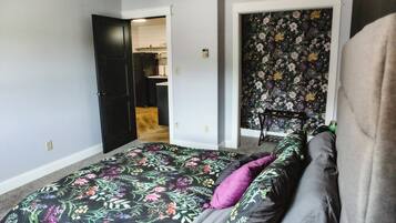 Ett sovrum med ett blommigt sängöverkast, en grå soffa och en dörr som leder in till ett kök.