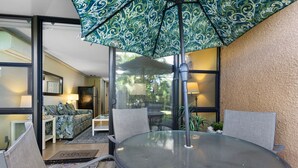A glass-enclosed patio with a round table, chairs, and a patterned umbrella. - Serene Hawaiian Beachside Bliss (Kihei)