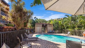 A swimming pool with lounge chairs and a covered area, surrounded by palm trees and a building with balconies. - Serene Hawaiian Beachside Bliss (Kihei)
