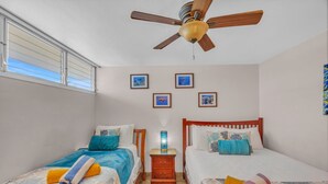 Une chambre avec deux lits simples, un ventilateur de plafond, une table de chevet avec une lampe et des tableaux encadrés au mur.