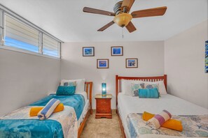 Un dormitorio con dos camas individuales, un ventilador de techo, una mesita de noche con lámpara y cuadros en la pared.