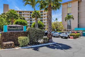 A hotel entrance with a sign reading "Kamaole Nalu," a parking lot with several cars, and a multi-story building in the background. - Oceanfront 2Bd 2Ba Air Conditioned Suite in South Maui (Kihei)