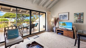 A spacious living area with a sliding glass door leading to a balcony, a flat-screen TV, and a wooden entertainment unit. - Oceanview Poolside Spacious 2-Bed Suite (Kihei)