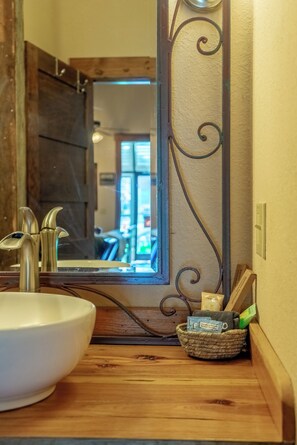 Un baño con tocador de madera, un lavabo blanco, un espejo con detalles decorativos en volutas y una cesta con artículos de aseo.