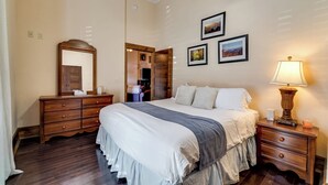 Un dormitorio con una cama grande, un aparador de madera y una mesita de noche con lámpara. Hay cuadros enmarcados en la pared y un espejo sobre el aparador.
