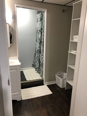 A small, narrow room with a shower, a white sink, a white rug, and a white laundry basket.