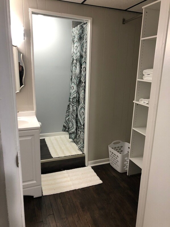 A small, narrow room with a shower, a white sink, a white rug, and a white laundry basket. - Studio Apartment on Main Street in Coshocton (25) (Coshocton)
