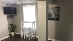 Una habitación con piso de madera, una mesa y sillas blancas, una planta en maceta, un televisor de pantalla plana montado en la pared y un cuadro en la pared.