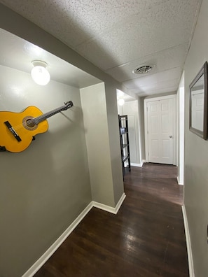 Een hal met een houten vloer, een gitaar die tegen de muur leunt, en een deur aan het einde.