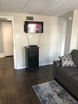 A living room with a flat-screen TV mounted on the wall, a dark wooden cabinet, a gray sofa with a patterned pillow, and a blue and white rug. - Modern apartment in downtown Coshocton (Coshocton)
