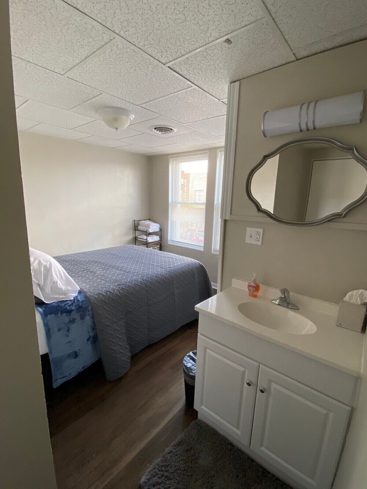 Une chambre d’hôtel avec un lit, une salle de bain comprenant un lavabo et un miroir, et une fenêtre avec des stores.