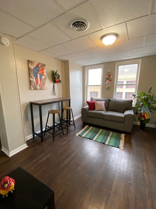 Una sala de estar con un sofá, una mesa de centro, un comedor con taburetes, un cuadro en la pared y un ventanal con vistas a edificios.