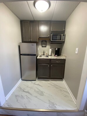 A compact kitchen with a stainless steel refrigerator, a microwave, dark wooden cabinets, a marble countertop, and a small sink.