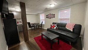 A compact living space with a black sofa, a red rug, a dining table with chairs, and a kitchenette in the background. - 1 Bedroom Apartment in Downtown Coshocton (23) (Coshocton)