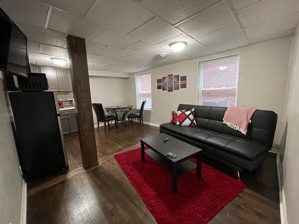 Ein gemütlicher Wohnbereich mit schwarzem Sofa, rotem Teppich, Esstisch mit Stühlen und einer kleinen Küchenzeile im Hintergrund.