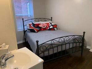 Ein Schlafzimmer mit einem Bettgestell aus Metall, einer grauen Bettdecke und Kissen mit dem Logo des Ohio State. Im Vordergrund ist ein weißes Waschbecken zu sehen.