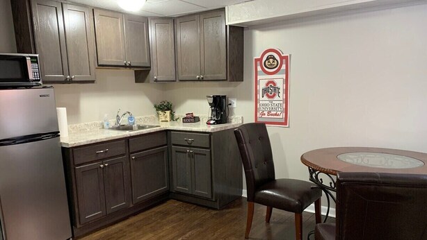 A compact kitchen with dark cabinets, a stainless steel refrigerator, a microwave, a coffee maker, and a dining table with chairs. - 1 Bedroom Apartment in Downtown Coshocton (23) (Coshocton)