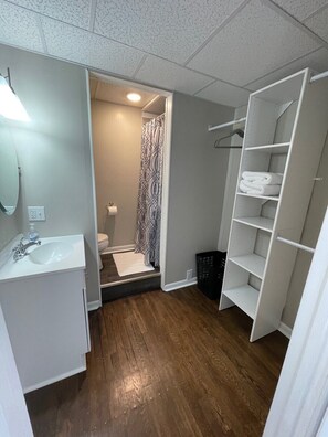 A bathroom with a white sink, toilet, and shower area. There is a white shelving unit and a black laundry basket. - Studio Apartment on Main Street in Coshocton (35) (Coshocton)