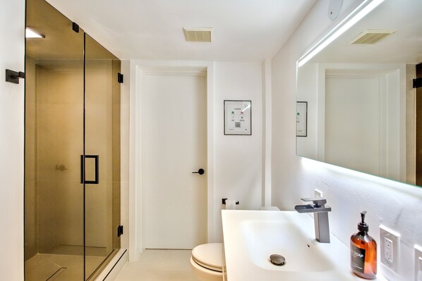 Un baño moderno con cabina de ducha de vidrio, inodoro blanco y lavamanos con dispensador de jabón.