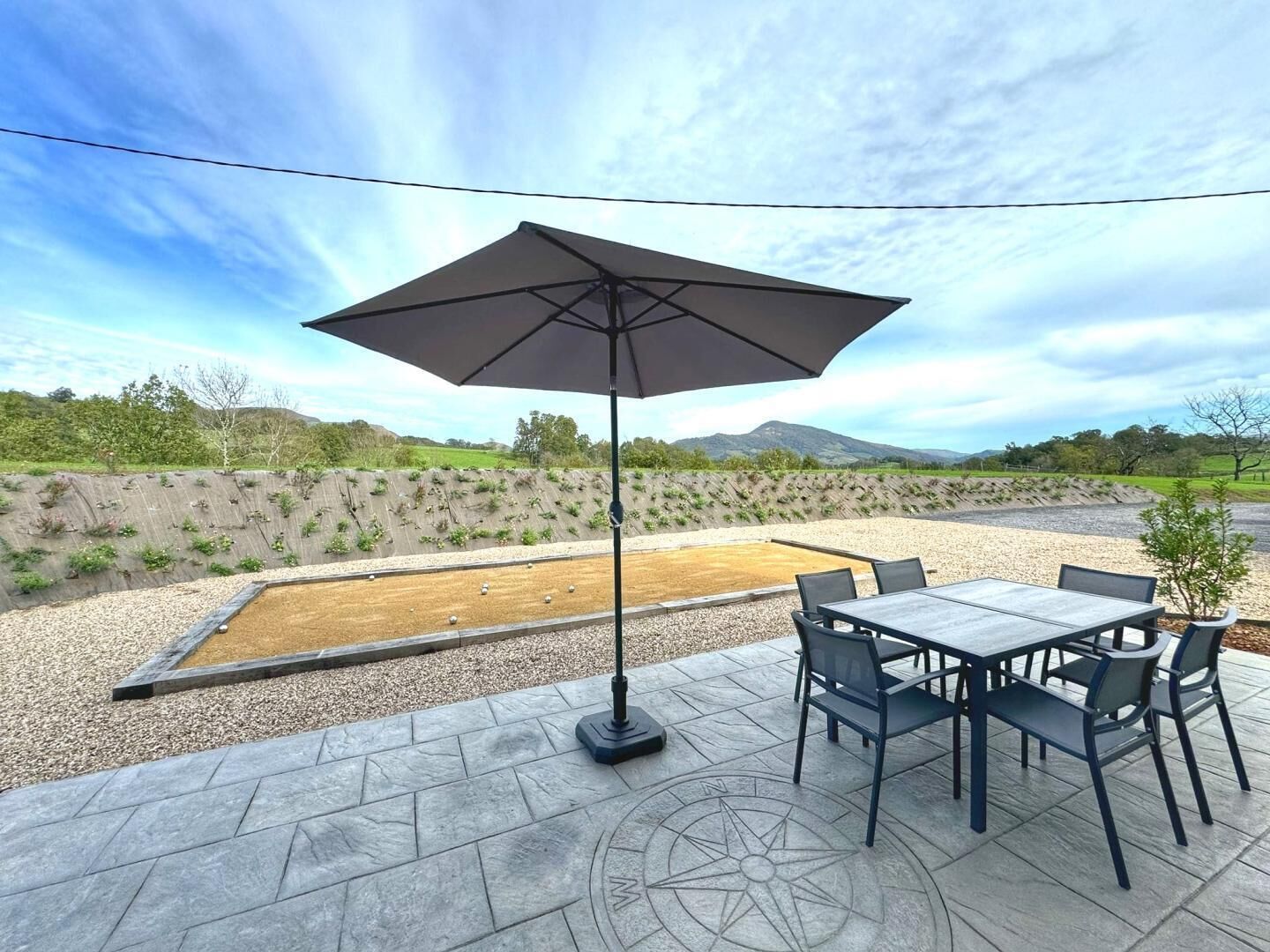 Un patio al aire libre con una mesa y sillas, un paraguas grande, y vistas a un área ajardinada con un muro de contención y colinas al fondo.