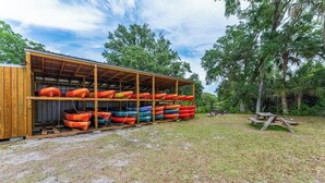 A covered boat storage area with multiple kayaks and a picnic table in a grassy outdoor setting. - Cozy River Cabin | Escape to Seven Sisters Springs (Homosassa)