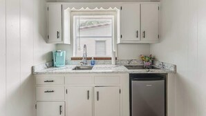 A compact kitchen with white cabinets, a stainless steel dishwasher, and a window with blinds. - Cozy River Cabin | Escape to Seven Sisters Springs (Homosassa)