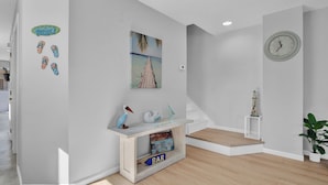 A modern, minimalist hallway with a wooden floor, a white console table with a 'BAR' sign, a painting of a beach, a clock, and a potted plant.