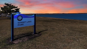 A sign for Sunset Lake Park with a view of the lake and a sunset in the background.