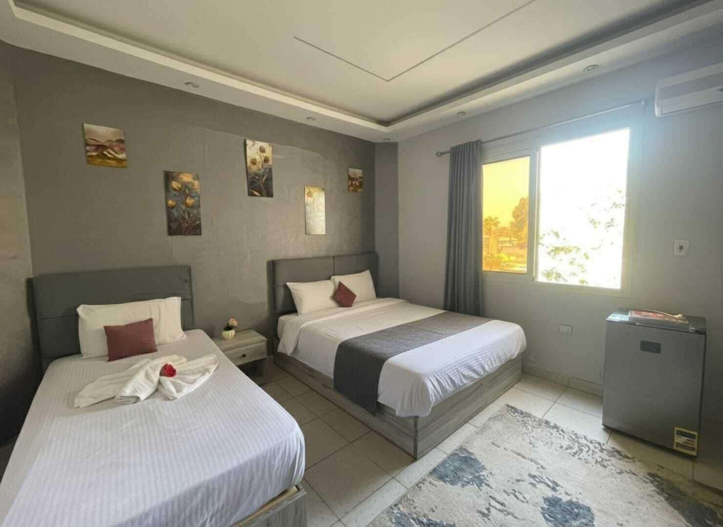 A hotel room with two beds, a small desk, a window with curtains, and artwork on the walls.