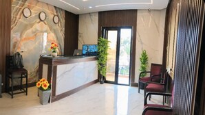 Ein Hotel-Empfangsbereich mit Marmor-Theke, zwei Uhren, einem Laptop und einer Sitzecke mit Stühlen und Topfpflanzen.