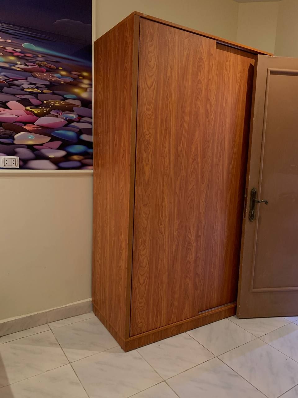 A wooden wardrobe with a handle, a tiled floor, and a wall with a colorful abstract painting.