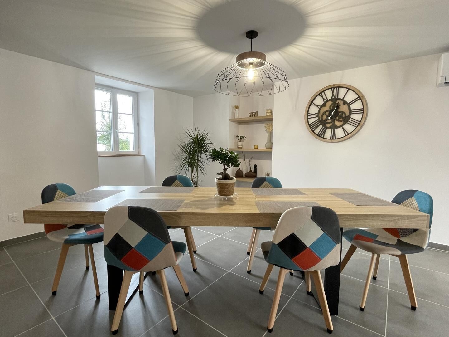 Un comedor con una gran mesa de madera, seis sillas con diseños geométricos, una lámpara colgante, un reloj de pared grande y una planta en maceta.