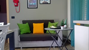 A living room with a grey sofa, green and yellow cushions, a small round table with a chess set, and two framed pictures on the wall.