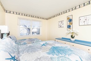 Quarto com cama, janela com cortinas de renda, mesa de cabeceira com um vaso de flores e decoração na parede.