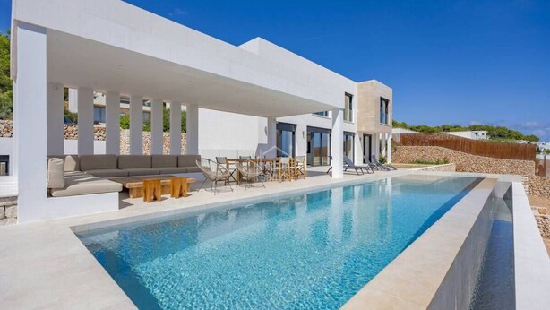 Un área de piscina moderna con una piscina cristalina, un espacio de descanso techado con sofá y sillones, y un muro de piedra al fondo.