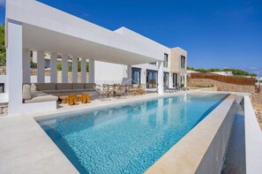 Un área de piscina moderna con una piscina cristalina, un espacio de descanso techado con sofá y sillones, y un muro de piedra al fondo.