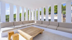 Un espacio exterior moderno con un amplio sofá seccional beige, una mesa de centro de madera y pilares blancos.
